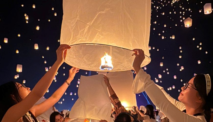 Một cặp đôi đang cùng nhau giữ và chuẩn bị thả một chiếc đèn lồng trong Lễ hội Yi Peng. Phía sau là nhiều chiếc đèn lồng khác đã được thả bay lên.