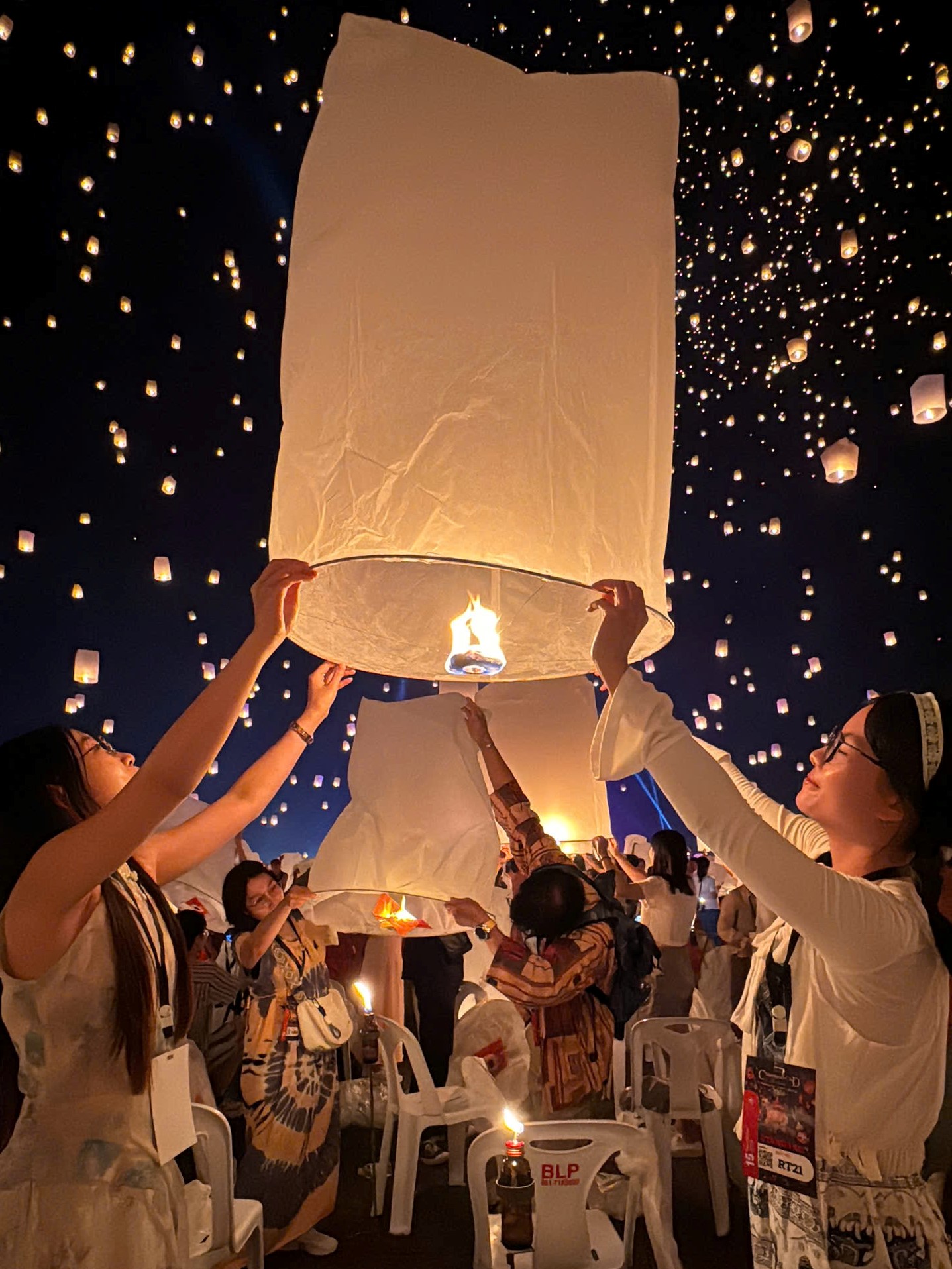 Một cặp đôi đang cùng nhau giữ và chuẩn bị thả một chiếc đèn lồng trong Lễ hội Yi Peng. Phía sau là nhiều chiếc đèn lồng khác đã được thả bay lên.