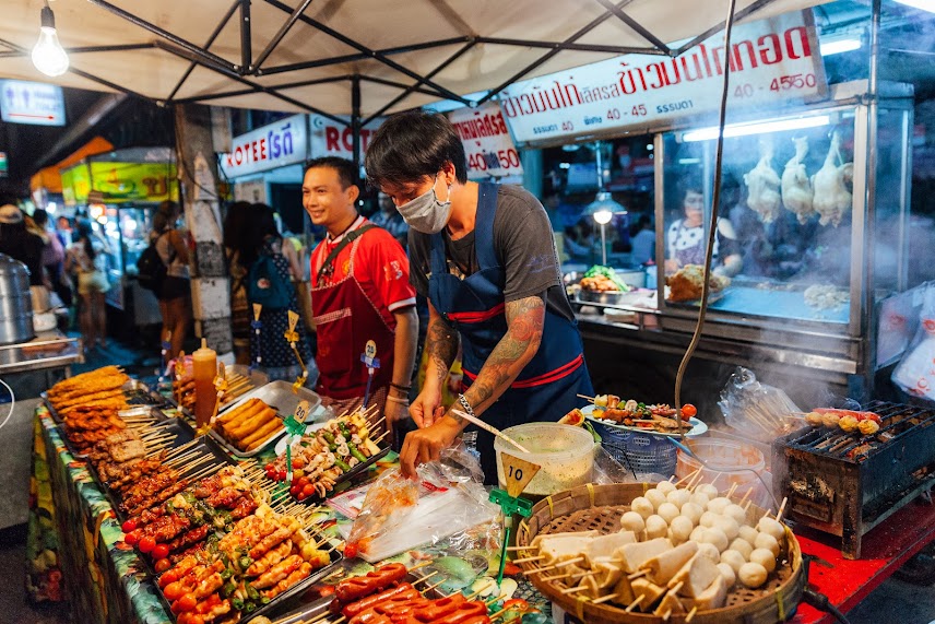 Một quầy hàng bán đồ ăn đường phố nhộn nhịp vào ban đêm ở Chiang Mai, Thái Lan, với người bán hàng đang chế biến các món xiên que nướng.