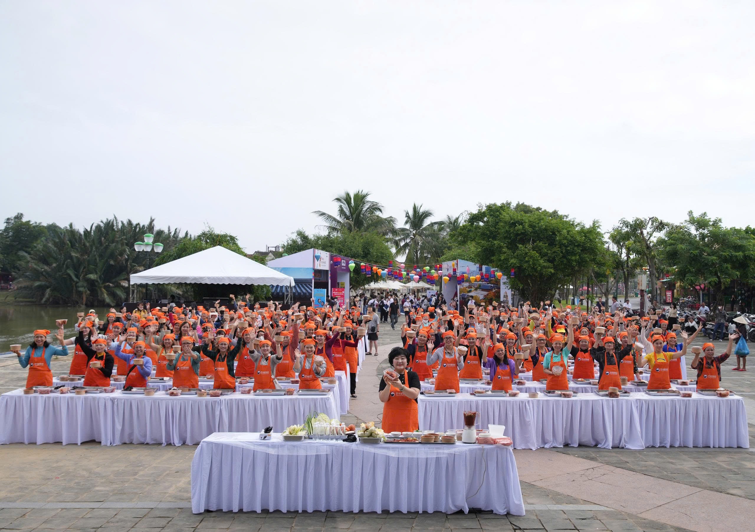 Toàn cảnh một nhóm đông người tham gia hoạt động làm kim chi, tất cả đều mặc tạp dề cam và đứng xung quanh các bàn dài.