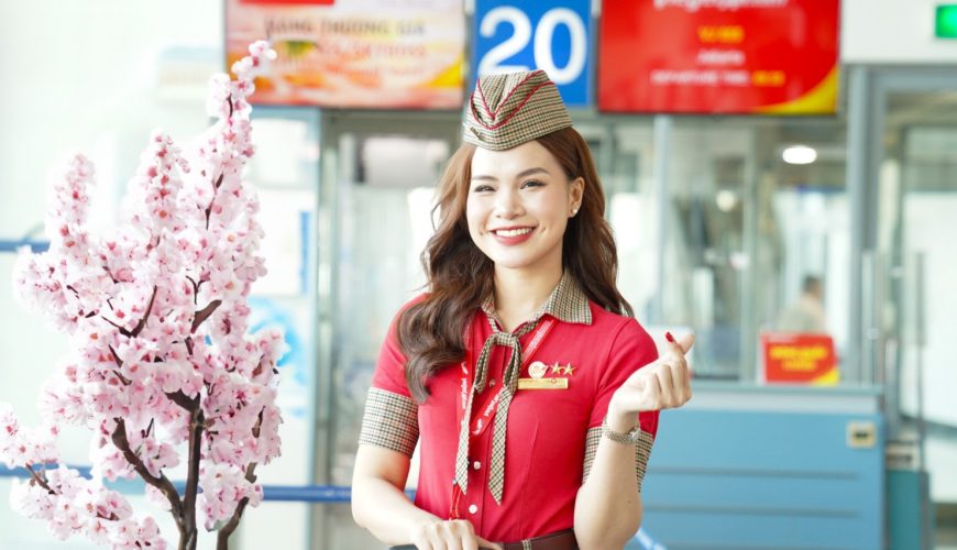 Nữ tiếp viên hàng không Vietjet tươi cười rạng rỡ, mặc đồng phục, đứng cạnh cây hoa anh đào giả tại sân bay.