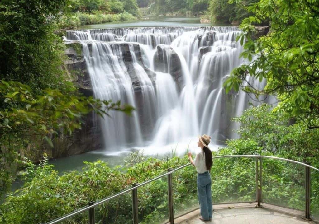 Cô gái đội mũ cói đứng trên đài quan sát bằng kính, ngắm nhìn Thác Nước Thập Phần hùng vĩ, bao quanh bởi rừng cây xanh tốt.
